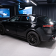 a prototype of the 2024 porsche macan electric photographed inside a convention center. we see the vehicle from the left-rear quarter view. it's painted black with vinyl wrapping to obscure some design details.
